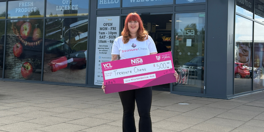 A woman standing in front of a Nisa Local storefront, holding a large ceremonial charity cheque for £500 made out to 